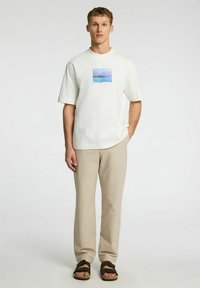 Vit kortärmad t-shirt med ett tryck av en blå och rosa landskap, tillsammans med beiga strukturerade byxor och bruna sandaler.