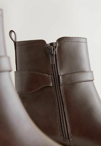 Bottines en cuir marron avec une fermeture éclair sur le côté, dotées d'une texture lisse et d'un détail de couture géométrique le long des coutures.