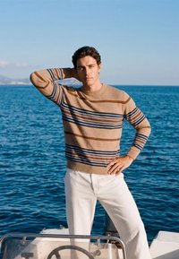 Homme en pull beige rayé et pantalon blanc, debout sur un bateau, une main sur la hanche et l'autre derrière la tête, avec la mer en arrière-plan.