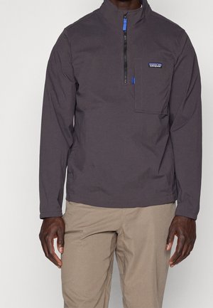 Homme portant un pull Patagonia gris foncé à demi-fermeture éclair avec une poche poitrine et un pantalon beige, debout devant un fond uni.