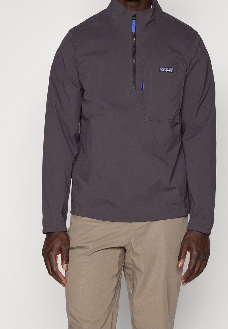 Homme portant un pull Patagonia gris foncé à demi-fermeture éclair avec une poche poitrine et un pantalon beige, debout devant un fond uni.