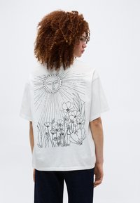 Γυναίκα με σγουρά μαλλιά, ντυμένη με λευκό πουκάμισο που έχει στο πίσω μέρος μια μαύρη γραφική παράσταση ενός ήλιου με πρόσωπο πάνω από άγρια λουλούδια.