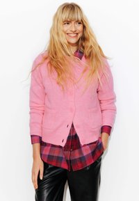 Cardigan rose en matériau doux, avec une fermeture à boutons et des poches. En dessous, une chemise à carreaux aux nuances de rouge et de bleu. Pantalons en cuir noirs.