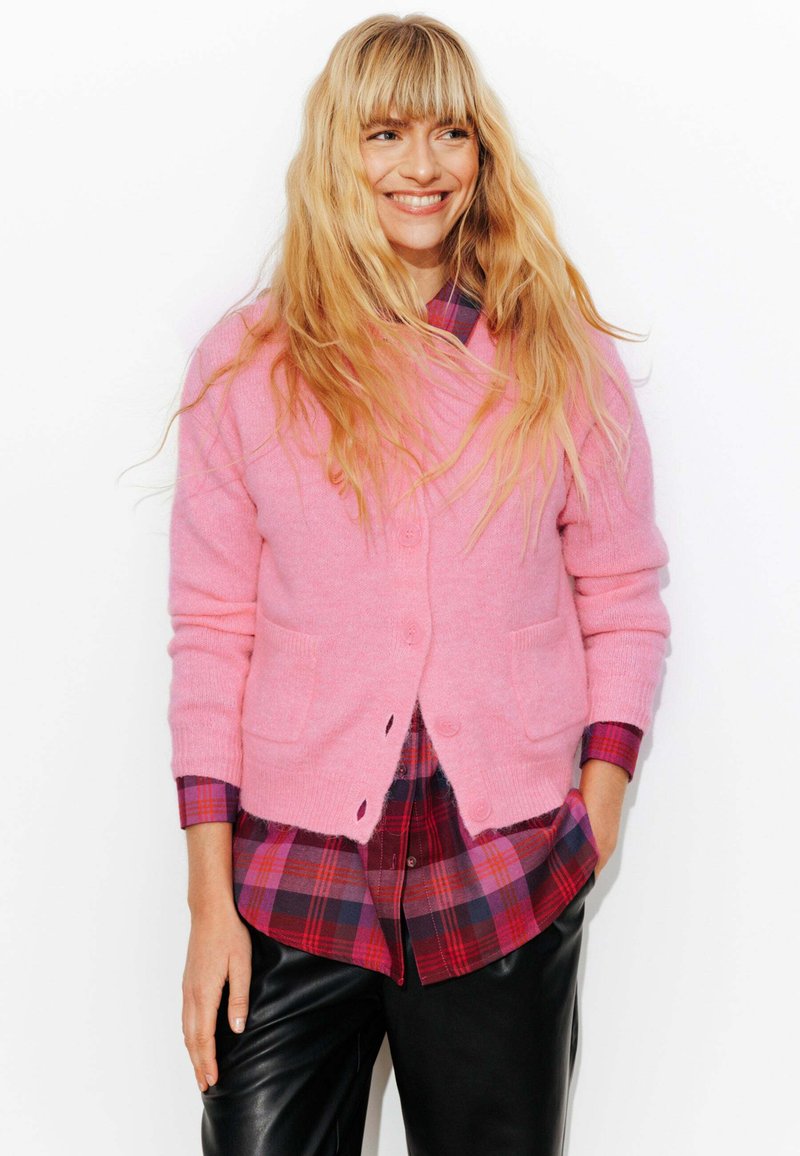 Cardigan rose en matériau doux, avec une fermeture à boutons et des poches. En dessous, une chemise à carreaux aux nuances de rouge et de bleu. Pantalons en cuir noirs.