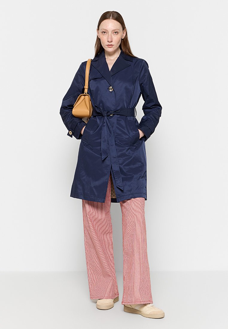 Trench coat blu navy con cintura in tessuto liscio, con collo e dettagli di bottoni, abbinato a pantaloni rossi a righe e calzature leggere.