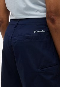 Άτομο που φοράει σορτς Columbia σε σκούρο μπλε χρώμα με ορατό λογότυπο της μάρκας στην πίσω τσέπη και ανοιχτόχρωμο πουκάμισο.