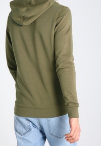 Felpa verde oliva con cappuccio regolabile, polsini e orlo a coste. Indossata sopra un jeans azzurro chiaro con cuciture visibili sui taschini posteriori.