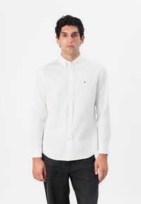 Camisa blanca de manga larga con botones hecha de algodón, con un cuello abotonado y un pequeño logo en el lado izquierdo del pecho. Se lleva con jeans oscuros.
