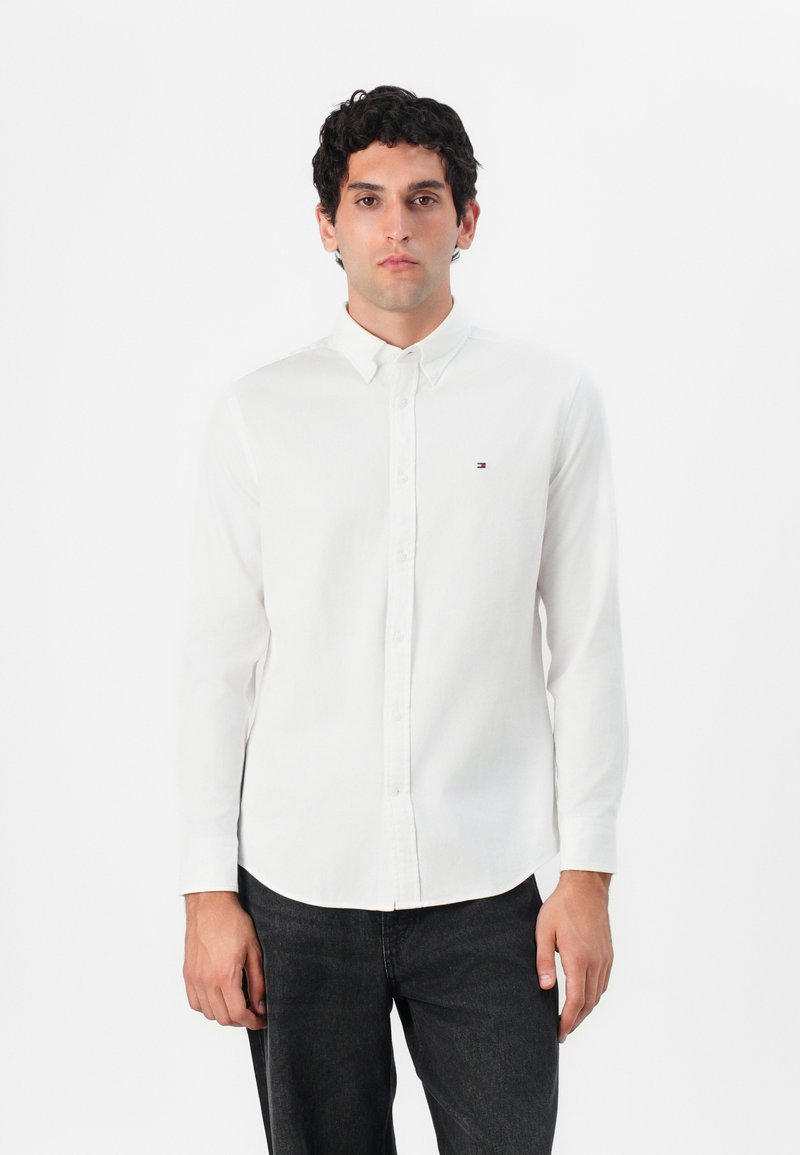 Camisa blanca de manga larga con botones hecha de algodón, con un cuello abotonado y un pequeño logo en el lado izquierdo del pecho. Se lleva con jeans oscuros.