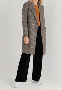 Donna che indossa un cappotto a quadri marrone, un maglione arancione, pantaloni larghi neri e sneakers bianche in piedi su uno sfondo neutro.