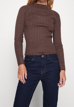 Brown ribbed long-sleeve sweater with a high neckline; fitted silhouette. Paired with dark blue high-waisted jeans showing curved hem.