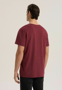 Mężczyzna o krótkich ciemnych włosach stoi tyłem, ubrany w jednolity bordowy t-shirt z krótkim rękawem i czarne spodnie na neutralnym tle.