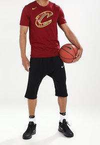 Camiseta de baloncesto roja de Nike con un logo amarillo en forma de "C", pantalones cortos negros, zapatillas negras y calcetines blancos. Sosteniendo un balón de baloncesto.