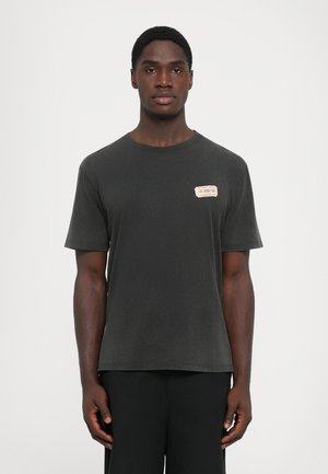 Mlad muž stoji, odjeven u jednostavnu tamno sivu majicu s malim bež zakrptom na lijevoj strani prsiju i crnim hlačama, na bijeloj pozadini.