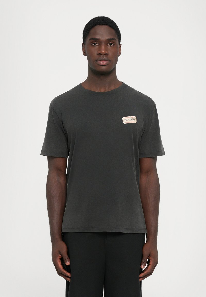 Jovem em pé, vestindo uma t-shirt cinza escura simples com um pequeno retângulo bege no lado esquerdo do peito e calças pretas, contra um fundo branco.