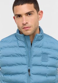 Junger Mann mit kurzen dunklen Haaren, der eine hellblaue gepolsterte Jacke mit hochgeklapptem Kragen und einem Markenpatch auf der Brust trägt.