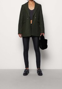 Blazer vert foncé à revers crantés et boutons doubles, associé à un pantalon noir ajusté. Le mannequin tient un sac à main noir et duveteux.