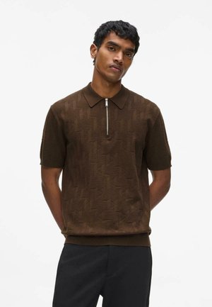 Rövid fekete hajú férfi, barna texturált rövid ujjú galléros pólót visel elején cipzárral és fekete nadrágot, kezeit a hát mögött tartva áll.