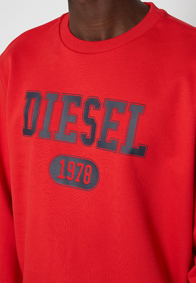 Persona che indossa una felpa rossa con la scritta "DIESEL" e "1978" stampate in grandi lettere grigie e rosse sul petto.