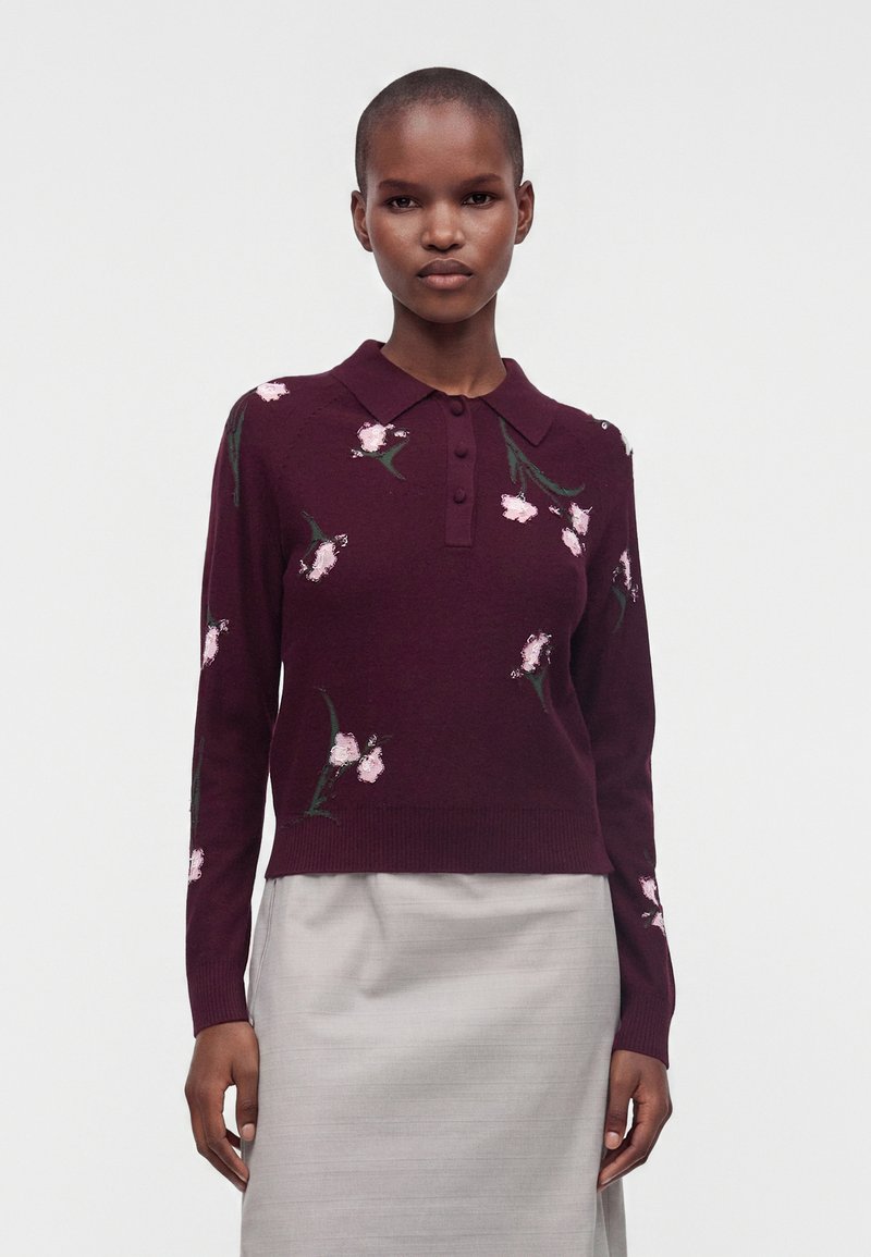 Frau steht und blickt nach vorne, trägt einen bordeauxfarbenen Blumensweater mit Kragen und Knöpfen sowie einen hellgrauen Rock vor einfarbigem Hintergrund.