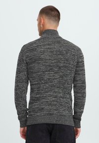 Hombre que lleva un suéter de cuello alto de punto jaspeado gris oscuro y pantalones negros, de espaldas a la cámara contra un fondo liso.
