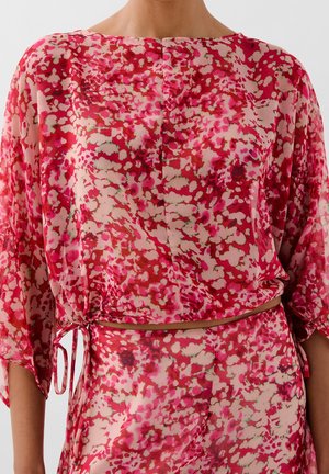 Close-up van een levendig roze en rood patroon blouse met losse mouwen en een trekkoord in de taille, gedragen met een bijpassende rok.