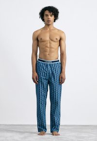 Lacoste blauwe, gemusterde pyjamabroek met een zwarte tailleband waarop de merknaam staat. Het materiaal lijkt zacht en heeft een geometrisch ontwerp.
