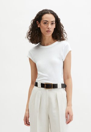 Kobieta o kręconych ciemnych włosach, ubrana w dopasowaną białą koszulkę z krótkim rękawem, jasnobeżowe spodnie z wysokim stanem oraz czarny pasek ze złotą sprzączką.