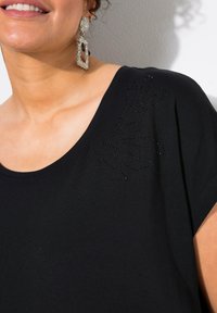 Czarna koszulka z okrągłym dekoltem, z teksturowanym wzorem i ozdobnymi koralikami na jednym ramieniu. Miękki, gładki materiał z krótkimi rękawami.