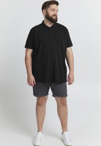 Černá polokošile s límečkem a třemi knoflíky, spárovaná s šedými šortkami. Model nosí bílé tenisky. Jednoduchý design, žádné viditelné vzory.