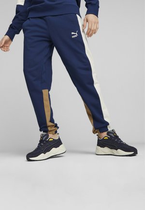 Pantalones de chándal azul marino con paneles laterales en crema y marrón, puños acanalados y un logo de Puma en blanco. Combinados con zapatillas oscuras con detalles en crema.