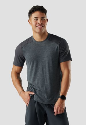 Lächelnder junger Mann, der ein dunkelgraues Sport-T-Shirt mit gemusterten Ärmeln, schwarze Shorts und eine schwarze Smartwatch trägt, mit einer Hand in der Tasche posiert.