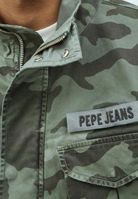 Maastokuvioinen takki hillityissä vihreän sävyissä, jossa on vetoketjukiinnitys, nepparikiinnitys, rintatasku ja kangasmerkki, jossa lukee "PEPE JEANS".