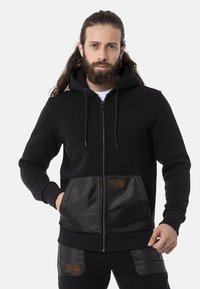 Schwarze Zip-up-Hoodie mit satinierter Textur, vorderer Tasche, Zugkordelkapuze, gerippten Bündchen und einem Lederlogo-Patch auf der Tasche.