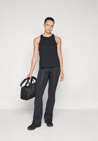Fekete atlétikai trikó széles pántokkal, fekete bővülő leggings-szel párosítva. Az alak egy kis fekete tornazsákot tart. Simaságú szövet textúra.