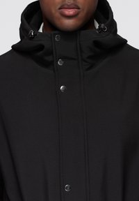 Czarna kurtka z kapturem wykonana z gładkiego materiału, z zatrzaskami i regulowanym sznurkiem przy kapturze, o minimalistycznym designie.