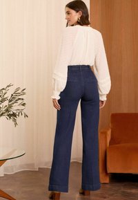 Uske strukirane, tamnoplave traperice visokog struka s širokim nogavicama. Ima stražnje džepove i manžete na rubovima. Nosive s kremastom bluzom.