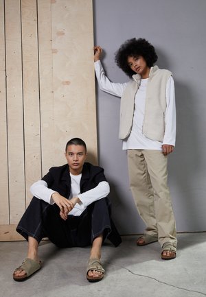 Twee modellen zijn gekleed in casual outfits. De ene draagt een beige fleece vest over een wit shirt, terwijl de andere een zwarte jas en wijde jeans heeft.