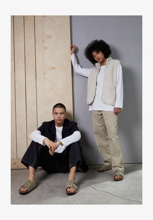 Twee modellen zijn gekleed in casual outfits. De ene draagt een beige fleece vest over een wit shirt, terwijl de andere een zwarte jas en wijde jeans heeft.