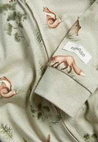 Zip-up beklædning i bomuld med en dæmpet beige baggrund og dyremotiver, herunder ræve og en bjørn, samt et mærke med teksten "newbie."