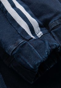 Jeansstoff in Dunkelblau mit weißem, gestreiftem Gummiband-Detail. Verfügt über einen gerafften Saum und eine glatte Textur. Fokus auf Naht und Verarbeitung.