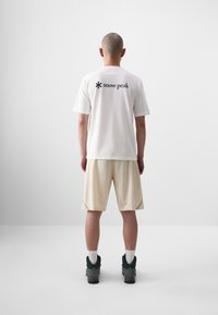 T-shirt blanche avec logo "snow peak" noir, shorts crème et chaussures foncées. Manches courtes, coupe décontractée, tissu lisse et détails de couture visibles.