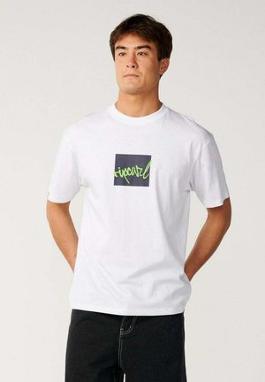 Homme portant un t-shirt blanc avec un carré noir et un logo en écriture verte, debout les mains derrière le dos sur un fond uni.