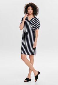 Vestido listrado verticalmente em azul-marinho e branco, com mangas curtas, decote em V, cinto removível e um corte descontraído. Sandálias escuras e finas completam o visual.