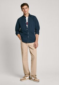 Camisa azul marino de botones sobre una camiseta gráfica blanca, pantalones beige y zapatillas color crema con acentos marrones. El modelo está de pie con una mano en el bolsillo.