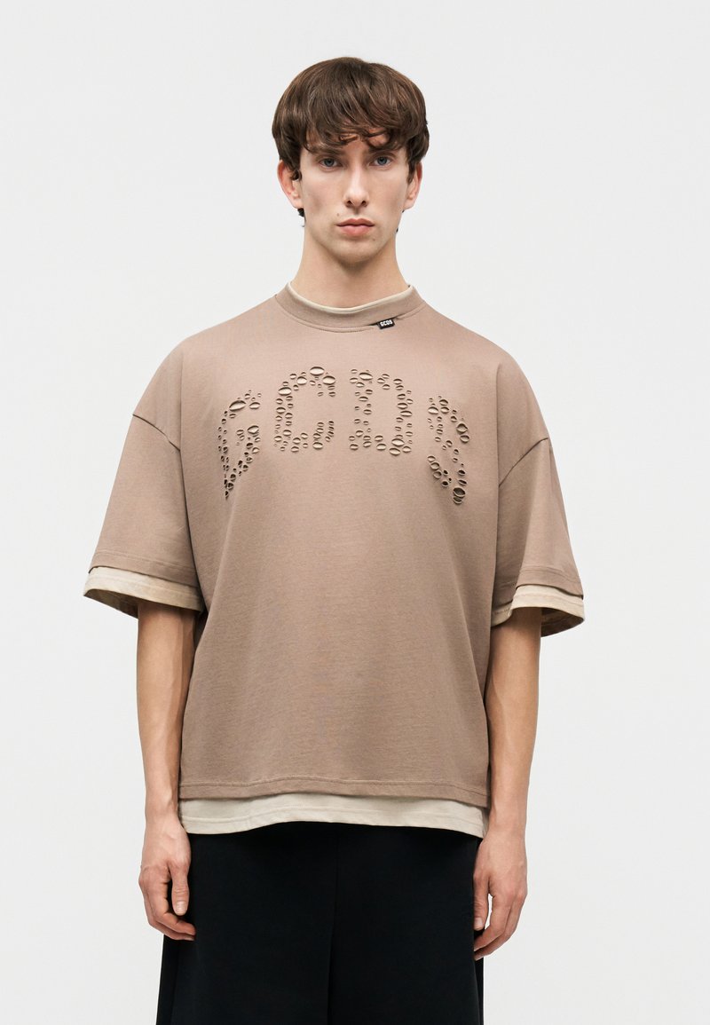 Νέος άνδρας με καστανά μαλλιά που φοράει μια μπεζ φαρδιά μπλούζα με κυκλικές οπές σχηματίζοντας γράμματα στο ύψος του στήθους, φορεμένη πάνω από ένα ανοιχτόχρωμο πουκάμισο.