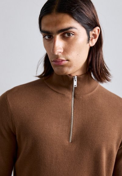 Junge Person mit langen dunklen Haaren, die einen braunen Reißverschluss-Pullover trägt, schaut direkt in die Kamera mit einem neutralen Gesichtsausdruck.