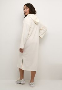 Langes Cream-Hoodie-Kleid aus weichem Stoff, mit langen Ärmeln, einem seitlichen Schlitz und einer lockeren Passform. Kombiniert mit hellen Sportschuhen.