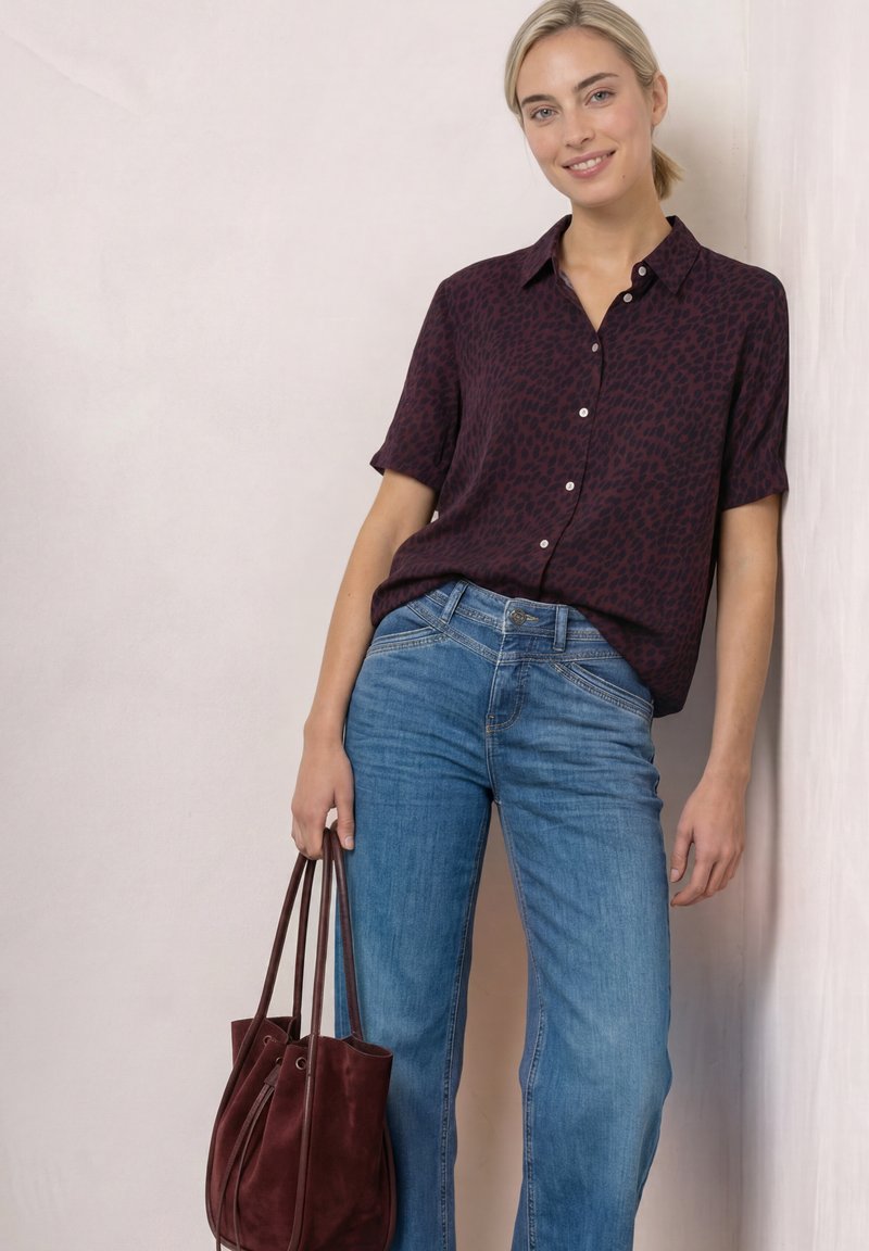 Femme portant une chemise à manches courtes foncée à motifs et un jean bleu, tenant un sac à main bordeaux, appuyée contre un mur clair, souriante.