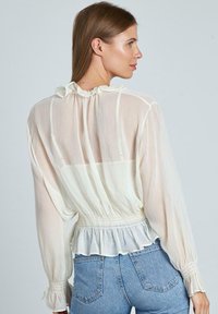 Witte, doorschijnende blouse met lange mouwen, rimpelnek en elastische taille. Heeft een gelaagd peplumdetail en verticale strepen.